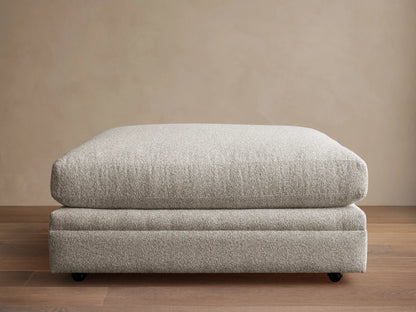 Rectangular Pavo Ottoman in light gray textured fabric with a plush top, on casters, resting on a wooden floor against a beige wall.