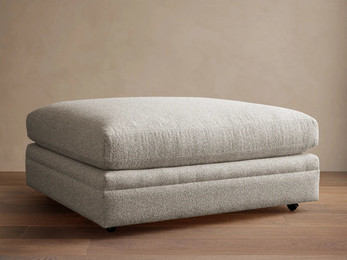 Rectangular Pavo Ottoman in light gray textured fabric with a plush top, on casters, resting on a wooden floor against a beige wall.