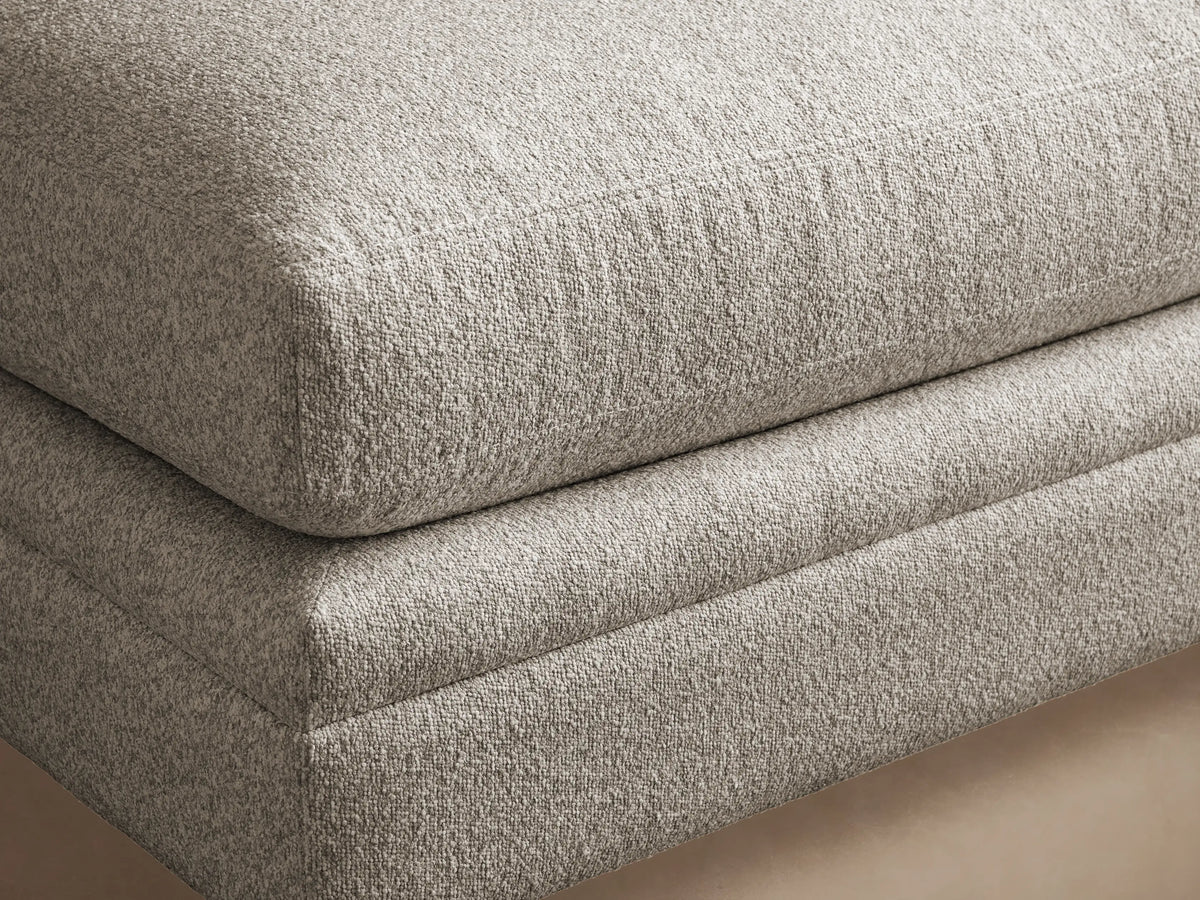Pavo Ottoman, light grey textured fabric, plush rectangular shape, on dark wood floor against a tan wall, viewed from front.