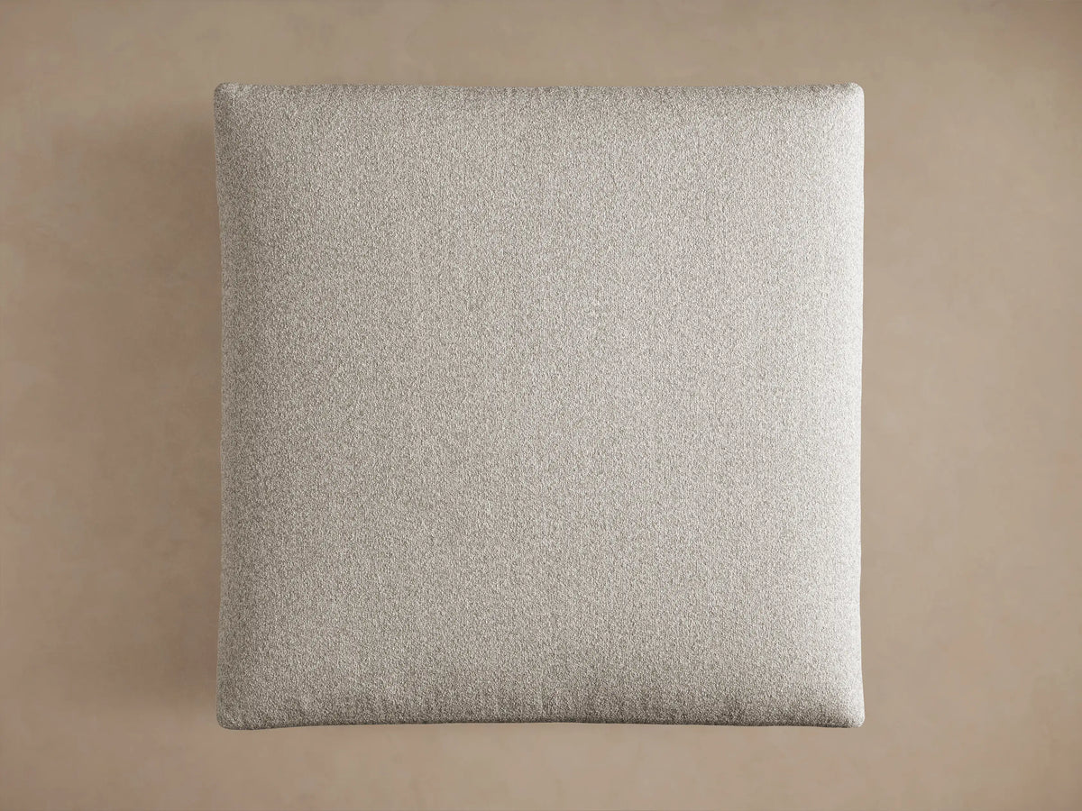 Close-up of a light gray, textured Pavo Ottoman, showing its plush top cushion resting on the upholstered base.