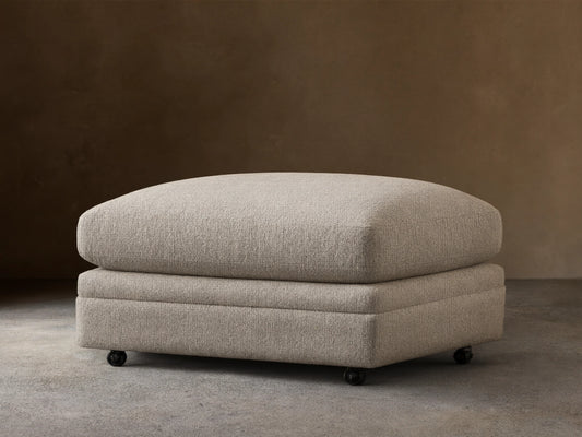 Rectangular Pavo Ottoman in light gray textured fabric with a plush top, on casters, resting on a wooden floor against a beige wall.