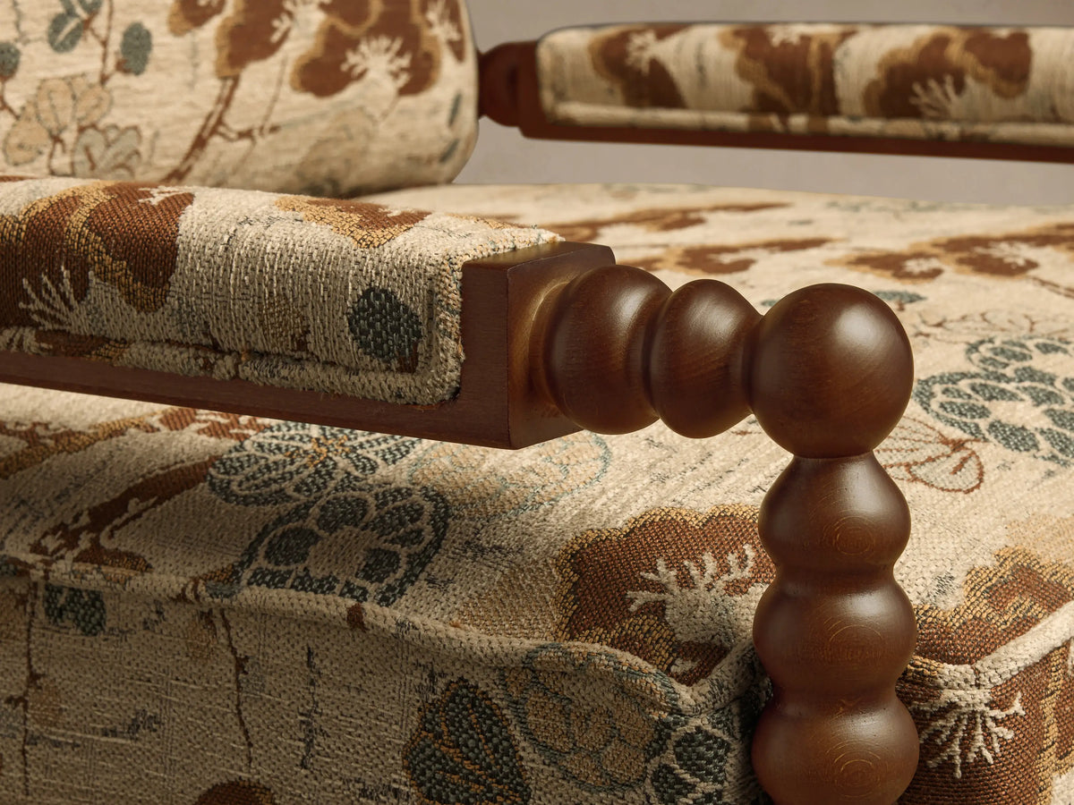 Close-up of the Milly Chair's armrest and side, showing light beige fabric with a brown and green floral pattern, and dark wood spindles.