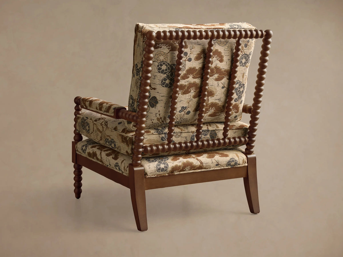 Rear 3/4 view of the Milly Chair. Dark wood frame with spindled details, and beige patterned upholstery with brown and blue florals.