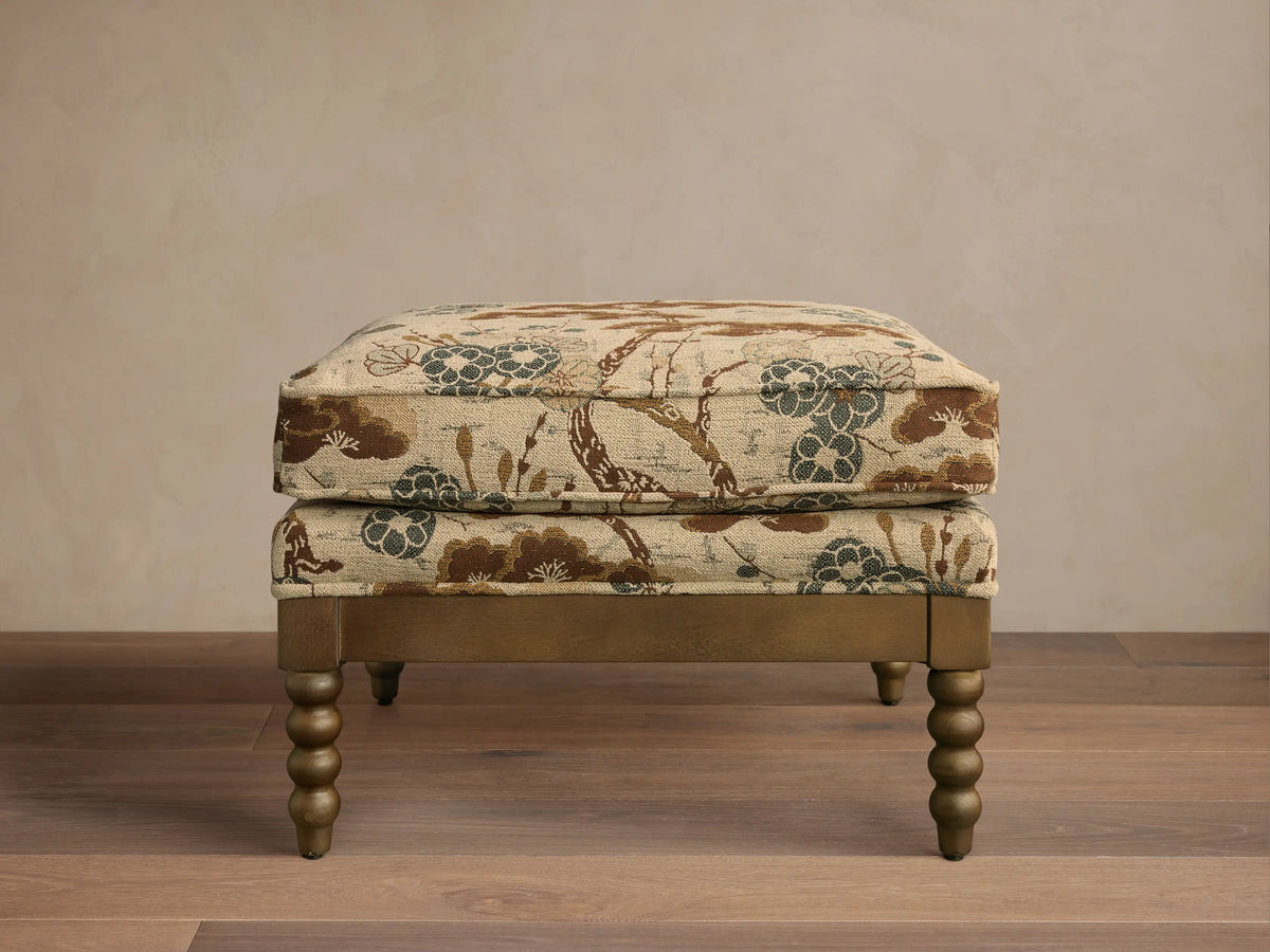 Front view of the Milly Ottoman, featuring beige fabric with brown, blue, and green floral pattern, and distressed gold spindled legs.