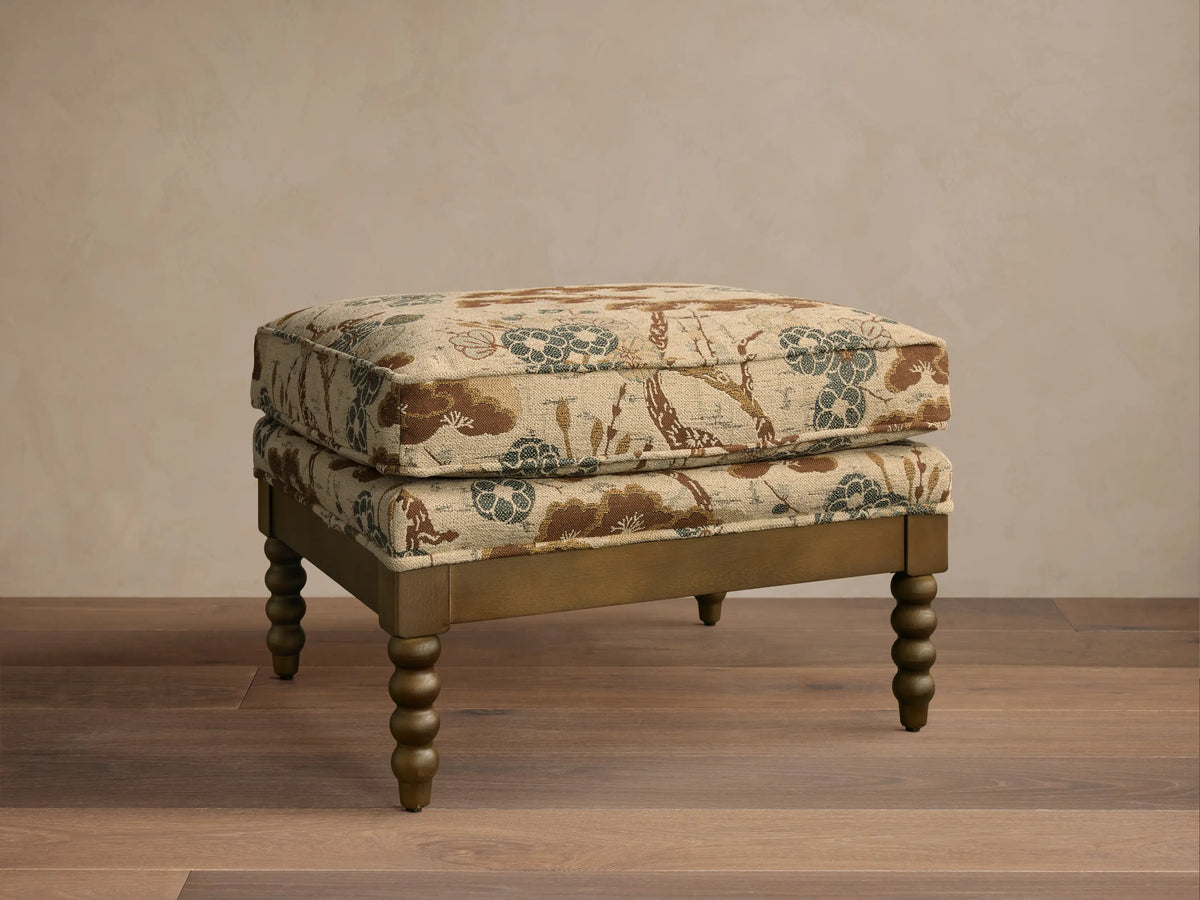 Front view of the Milly Ottoman, featuring beige fabric with brown, blue, and green floral pattern, and distressed gold spindled legs.