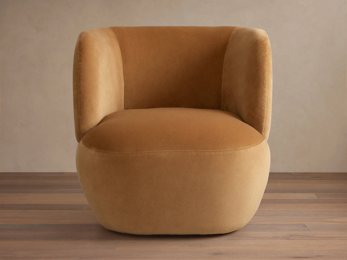 Lenny Swivel Chair in caramel brown fabric with a rounded seat, curved backrest, stout arms, and smooth upholstery on wooden floor.