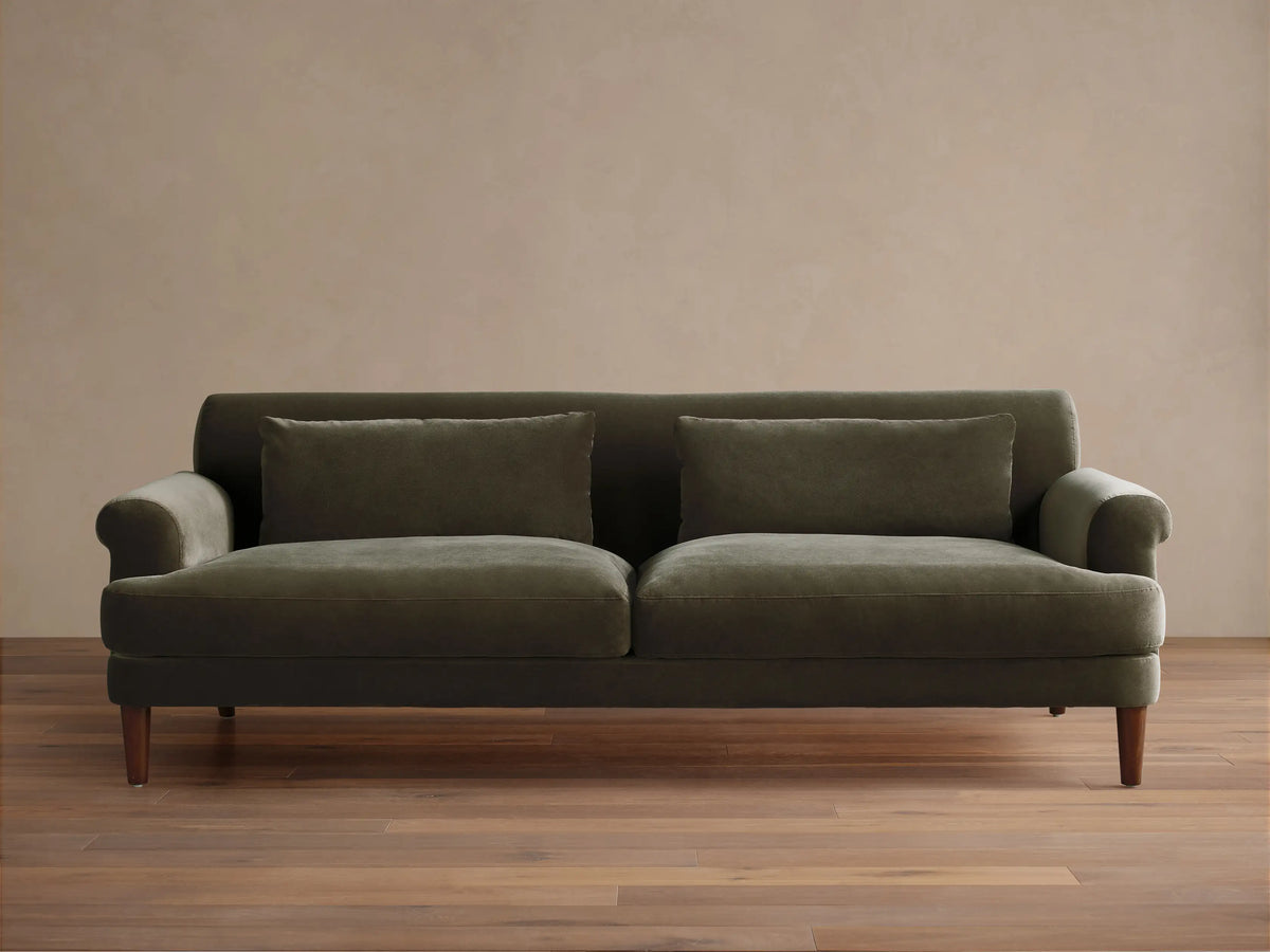 Thorne Sofa in dark olive green velvet, front view, with two seat cushions, two back pillows, and dark wood legs, on a wood floor.