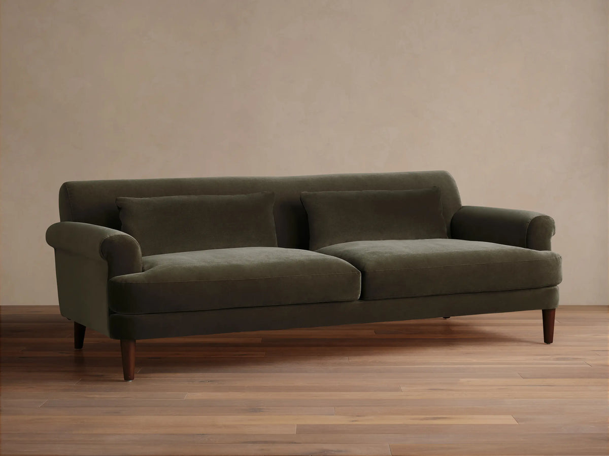 Thorne Sofa in dark olive green velvet, front view, with two seat cushions, two back pillows, and dark wood legs, on a wood floor.