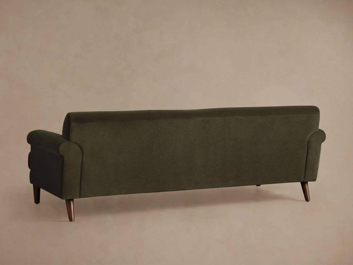 Angled view of the Thorne Sofa. Dark olive green velvet upholstery, featuring a rounded armrest, seat cushions, and dark wooden leg.