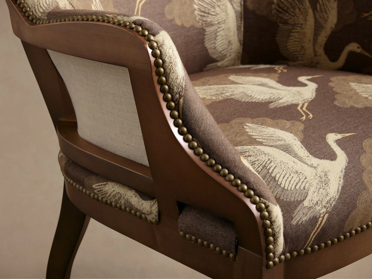 Front view of the brown Tilly Chair with beige crane and cloud pattern upholstery, dark wooden legs, and brass nailhead trim.