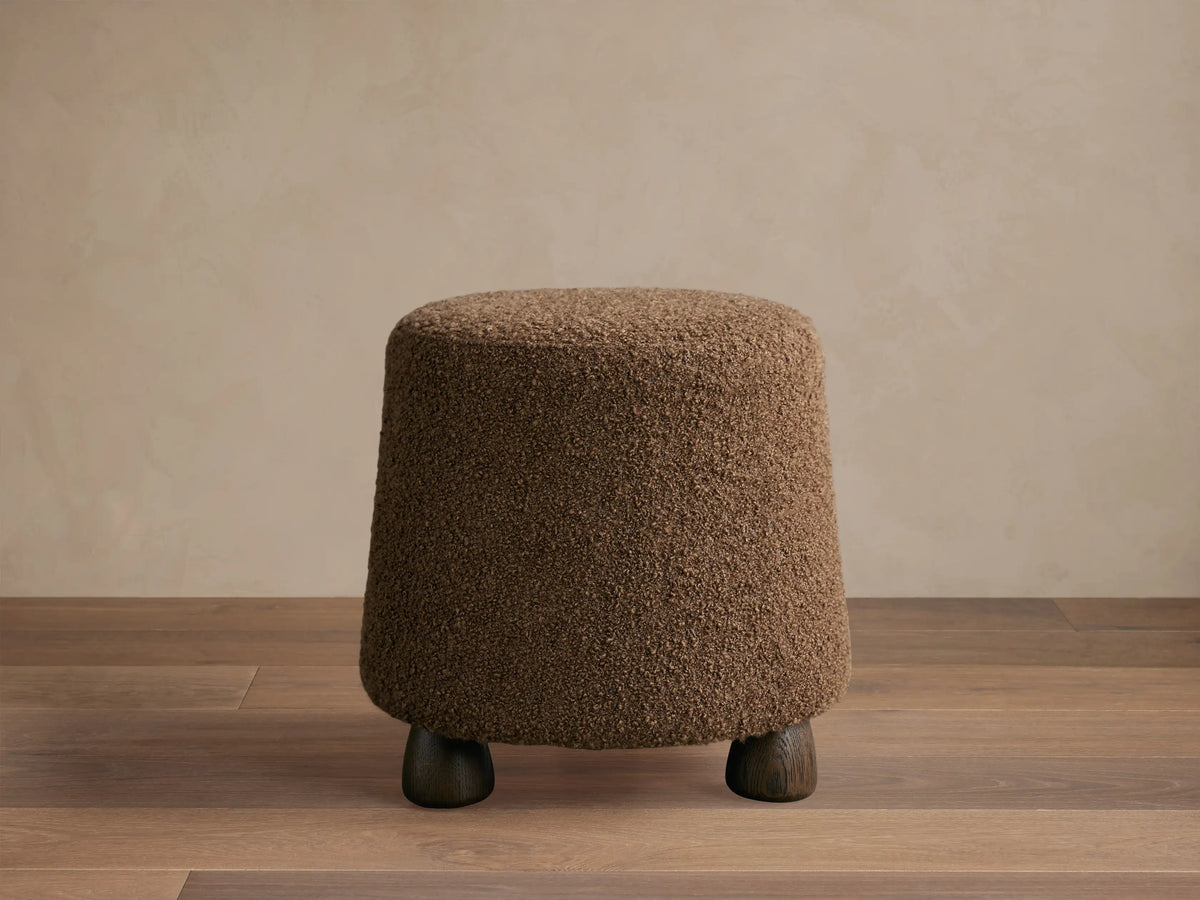 Steffi Ottoman, a brown, tapered bouclé-textured ottoman with three dark chestnut wood legs on a wooden floor.
