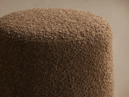 Brown textured Steffi Ottoman, round with chestnut wooden legs, positioned next to a beige bed on a patterned rug.