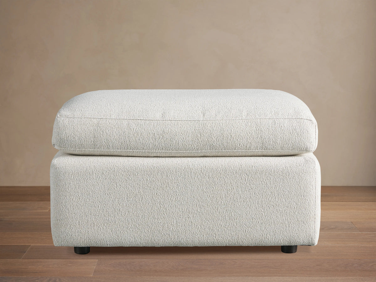 Ashby Ottoman in light cream CryptonÂ® fabric featuring a rectangular cushion on a solid base with black feet, set on a wooden floor.