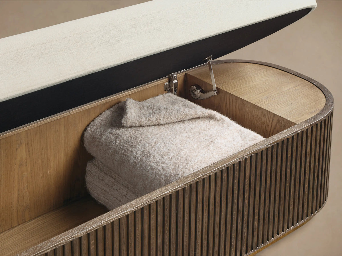 Front view of the oval Finnley Storage Bench with a cream upholstered top and dark fluted wood base with bronze trim.