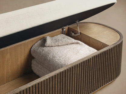 Front view of the oval Finnley Storage Bench with a cream upholstered top and dark fluted wood base with bronze trim.