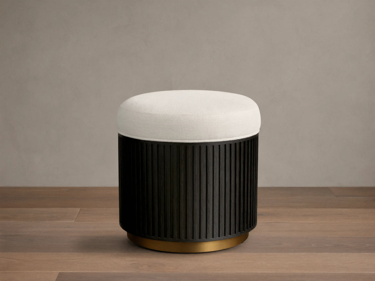 Front view of the Finnley Ottoman featuring a cream upholstered cushion, an Ebony vertical ribbed wood base, and a brass-colored circular trim.
