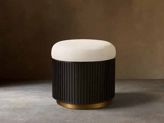 Front view of the Finnley Ottoman featuring a cream upholstered cushion, an Ebony vertical ribbed wood base, and a brass-colored circular trim.