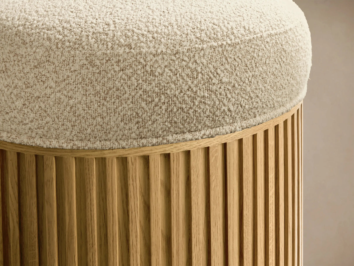 Front view of the round Finnley Ottoman, featuring a light textured cushion, slatted honey-finished wood base, and metallic ring.