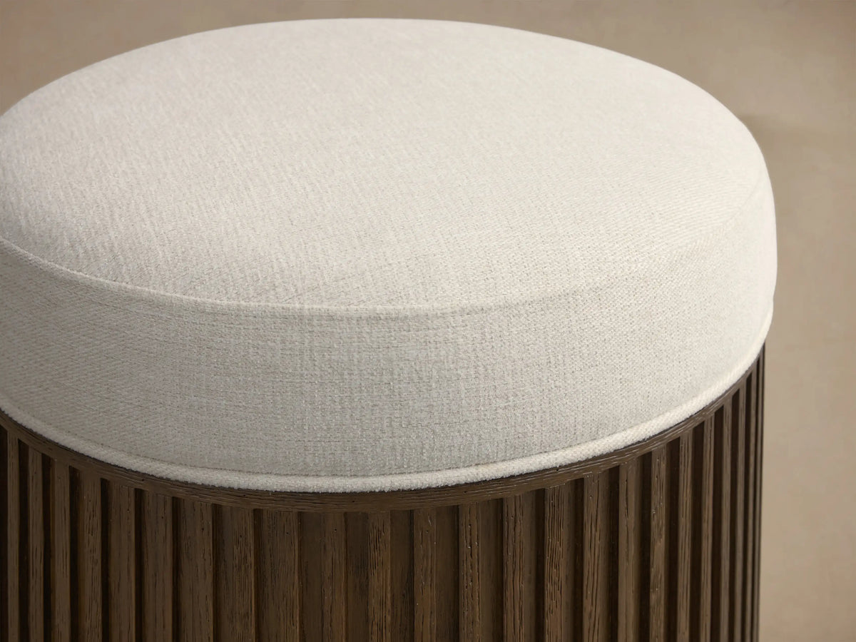 Front view of the round Finnley Ottoman featuring a cream upholstered top and dark wood fluted base with a golden trim.