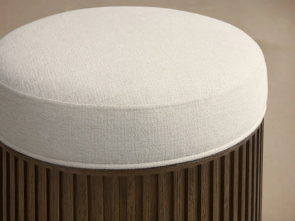 Front view of the round Finnley Ottoman featuring a cream upholstered top and dark wood fluted base with a golden trim.