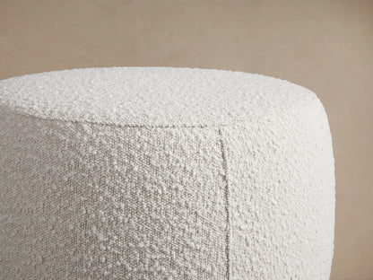 Round, textured grey Ryles Ottoman on wooden floor. The fabric has a nubby appearance. Plain background highlights the ottoman's minimalist design.