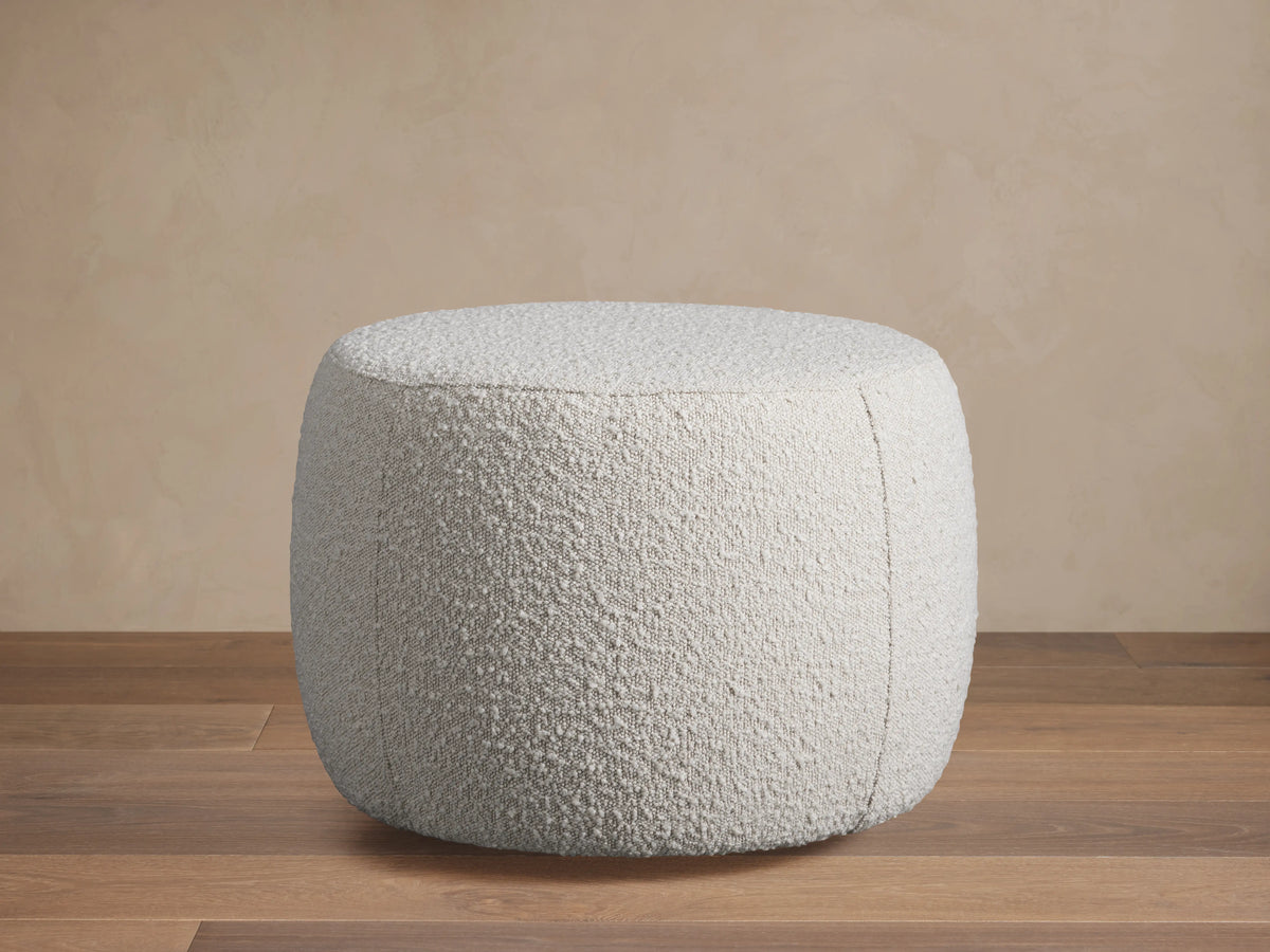 Ryles Ottoman in textured white fabric, beside a matching chair on a wooden floor, set against a beige wall background.