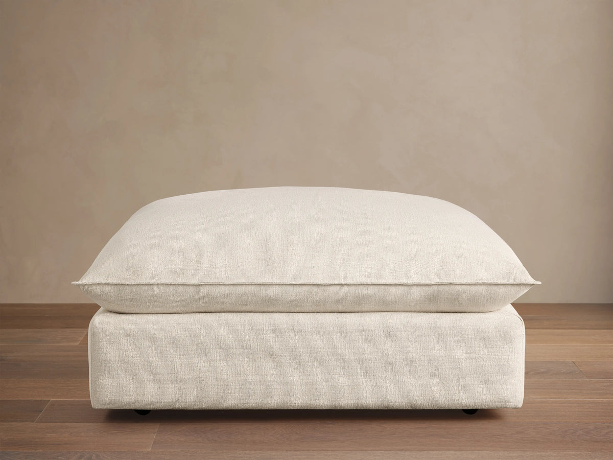 Ottoman with a soft, off-white cushioned top and a matching base, set against a wooden floor and beige wall, featuring subtle caster wheels.