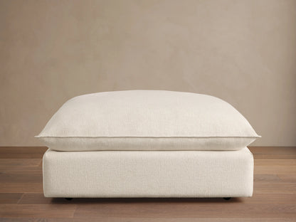 Ottoman with a soft, off-white cushioned top and a matching base, set against a wooden floor and beige wall, featuring subtle caster wheels.