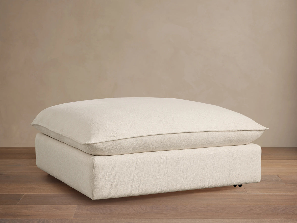 Ottoman with a soft, off-white cushioned top and a matching base, set against a wooden floor and beige wall, featuring subtle caster wheels.