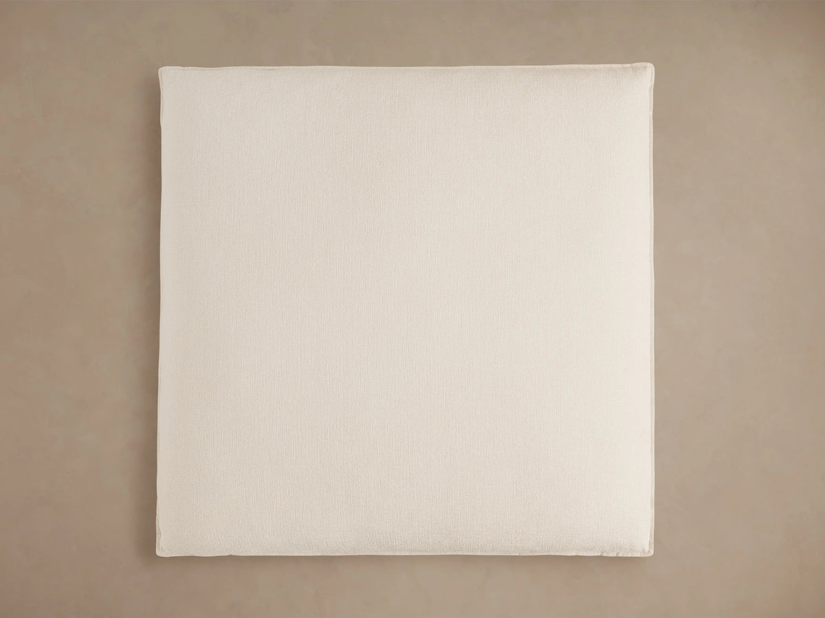 Owen Ottoman in close-up, showcasing beige textured fabric with visible stitching, featuring a plush cushion on top, and blurred neutral background.