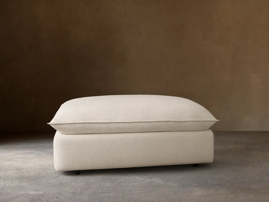 Ottoman with a soft, off-white cushioned top and a matching base, set against a wooden floor and beige wall, featuring subtle caster wheels.