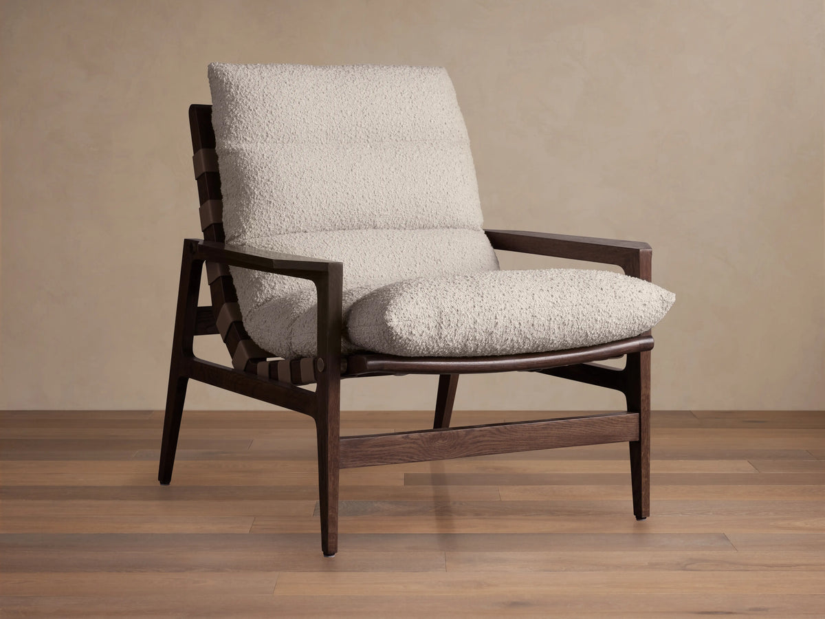 Rourke Chair with solid hardwood frame, neutral sand bouclé cushions, leather straps, and brass accents on a wood floor.