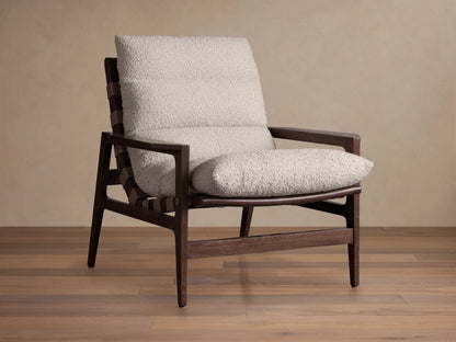 Rourke Chair with solid hardwood frame, neutral sand bouclé cushions, leather straps, and brass accents on a wood floor.