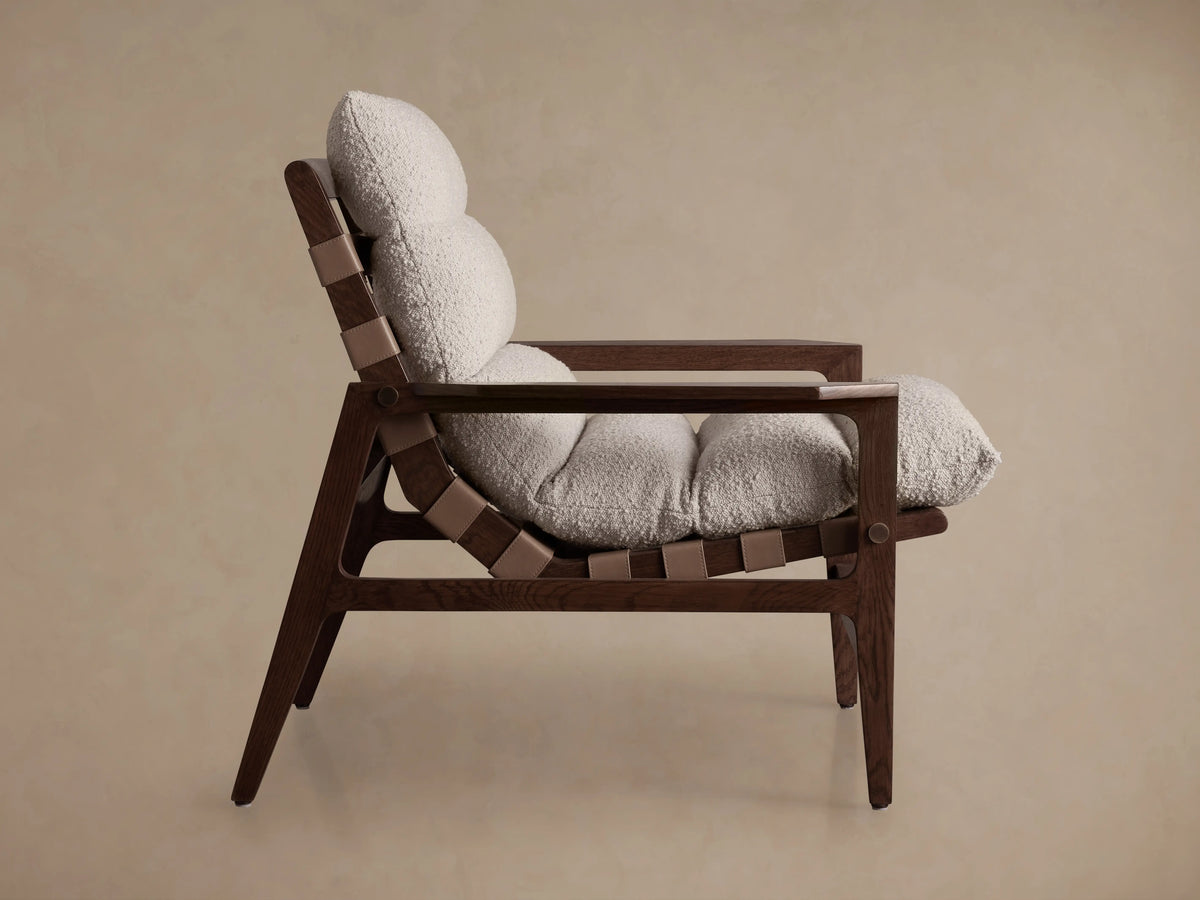 Rourke Chair side view with a dark wood frame, textured sand-colored bouclé cushions, and leather straps with brass accents.
