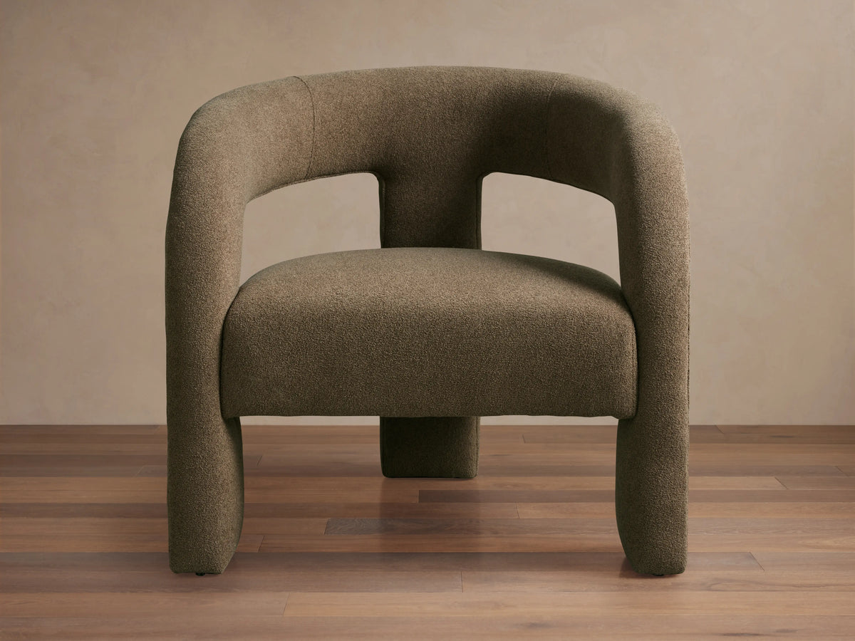 Mara Cutout Chair in taupe fabric, showcasing a rounded backrest and cutout design. Positioned on a wooden floor.