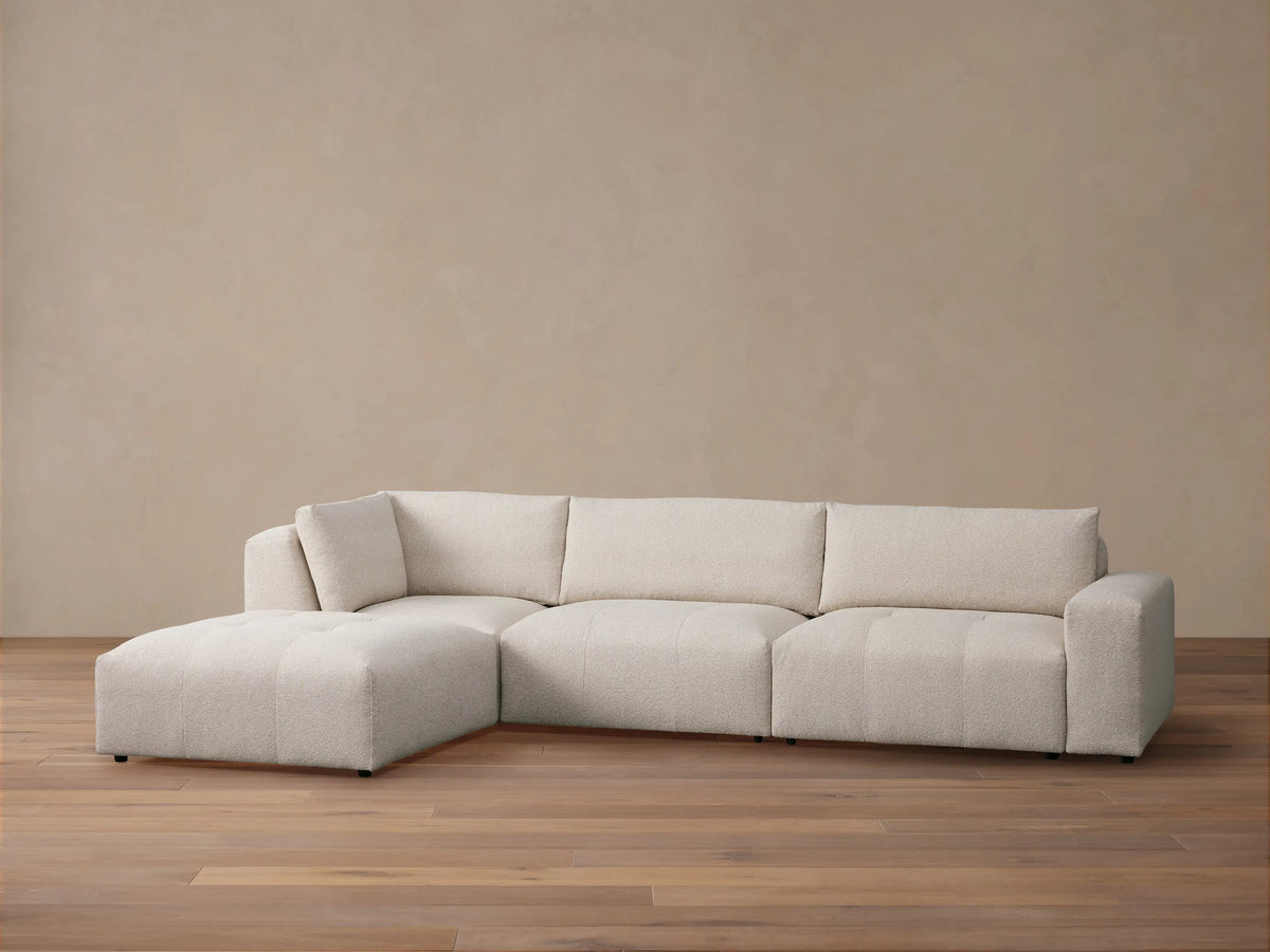 Cream Keegan L-shaped sectional sofa with ottoman on left, on a wooden floor against a light beige wall.