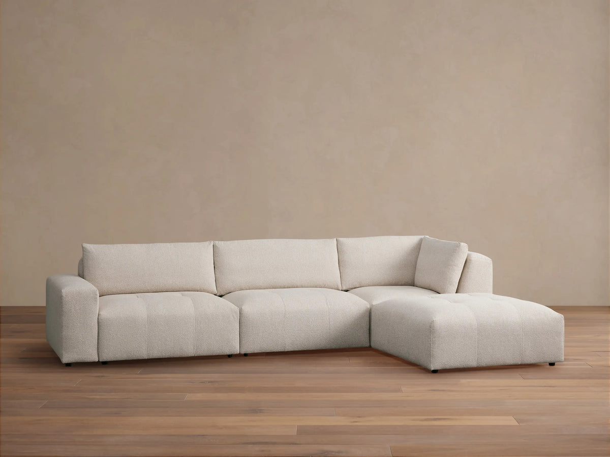 Keegan Four Piece Sectional with Ottoman in light beige textured fabric, L-shaped, on a brown wood floor.