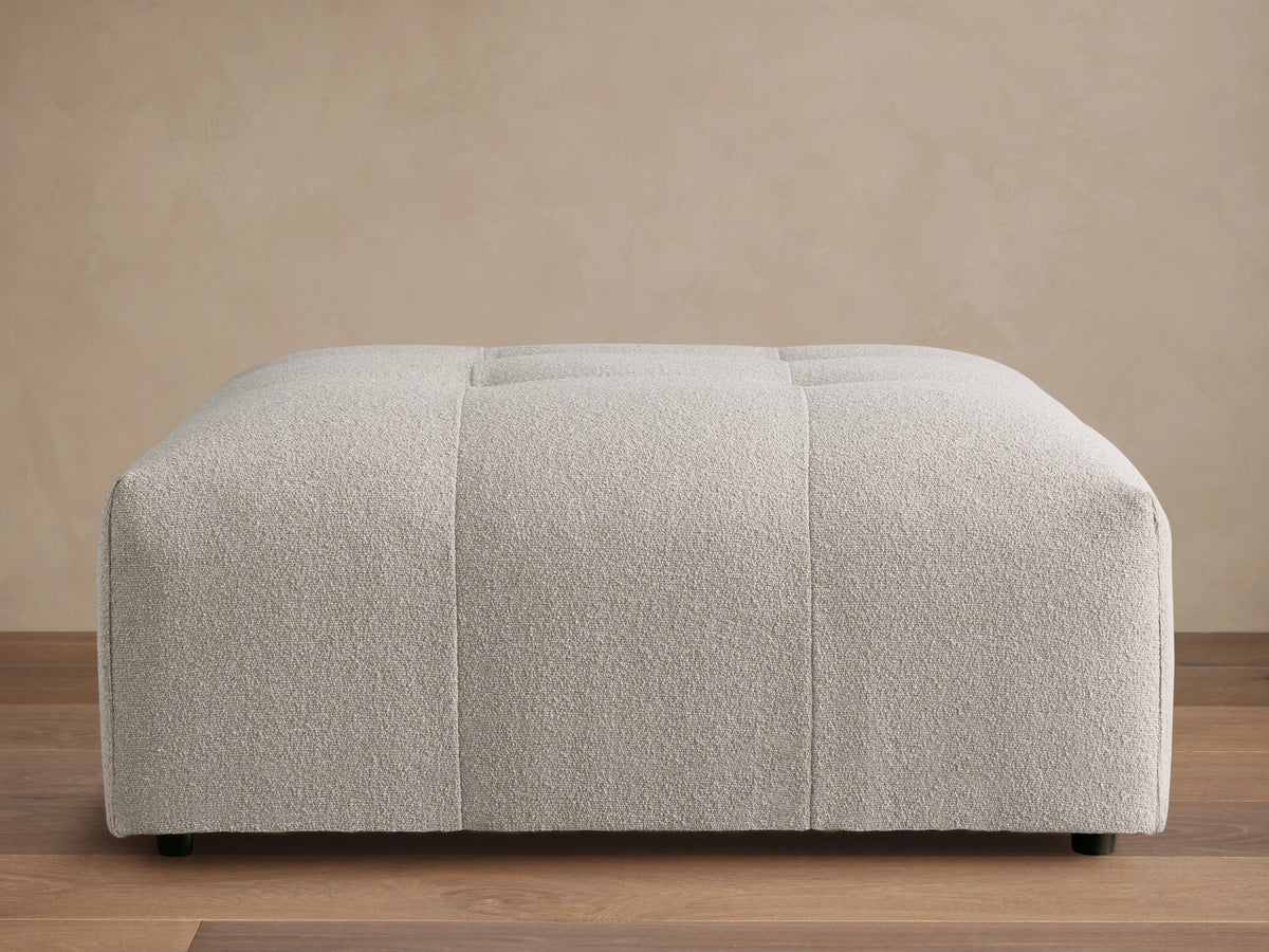 Keegan Ottoman in light grey textured fabric with subtle tufting on top, resting on a wooden floor against a beige wall.