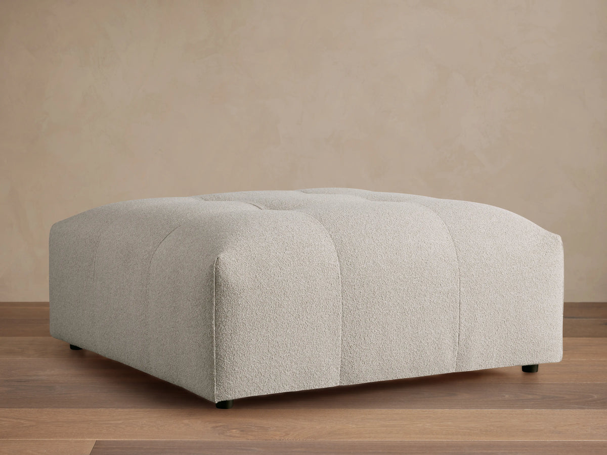 Keegan Ottoman in light grey textured fabric with subtle tufting on top, resting on a wooden floor against a beige wall.