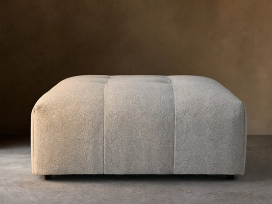 Keegan Ottoman in light grey textured fabric with subtle tufting on top, resting on a wooden floor against a beige wall.