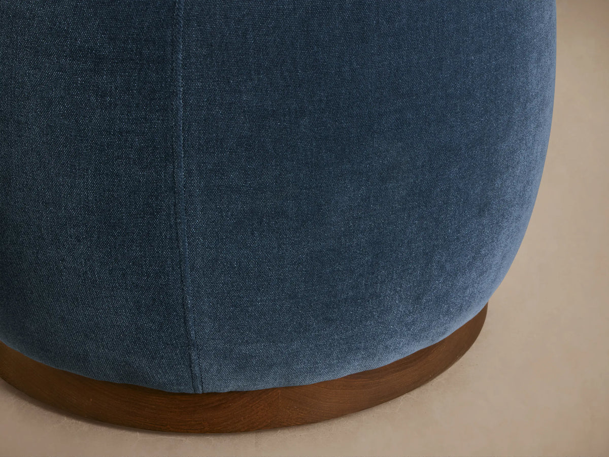 Close-up of Ryles Plinth Ottoman's lower section: deep blue textured fabric with a vertical seam, resting on a round brown wooden base.