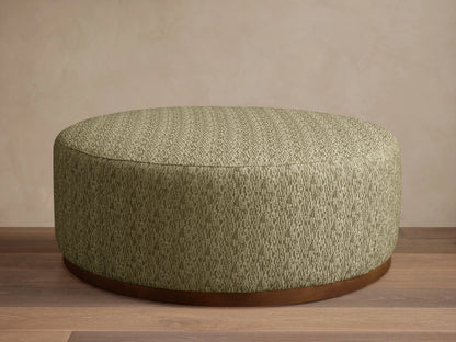 Round Ryles Plinth Ottoman, upholstered in textured light olive green fabric with a dark wood base, on a wooden floor.