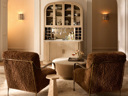 Round Ryles Plinth Ottoman, upholstered in textured light olive green fabric with a dark wood base, on a wooden floor.