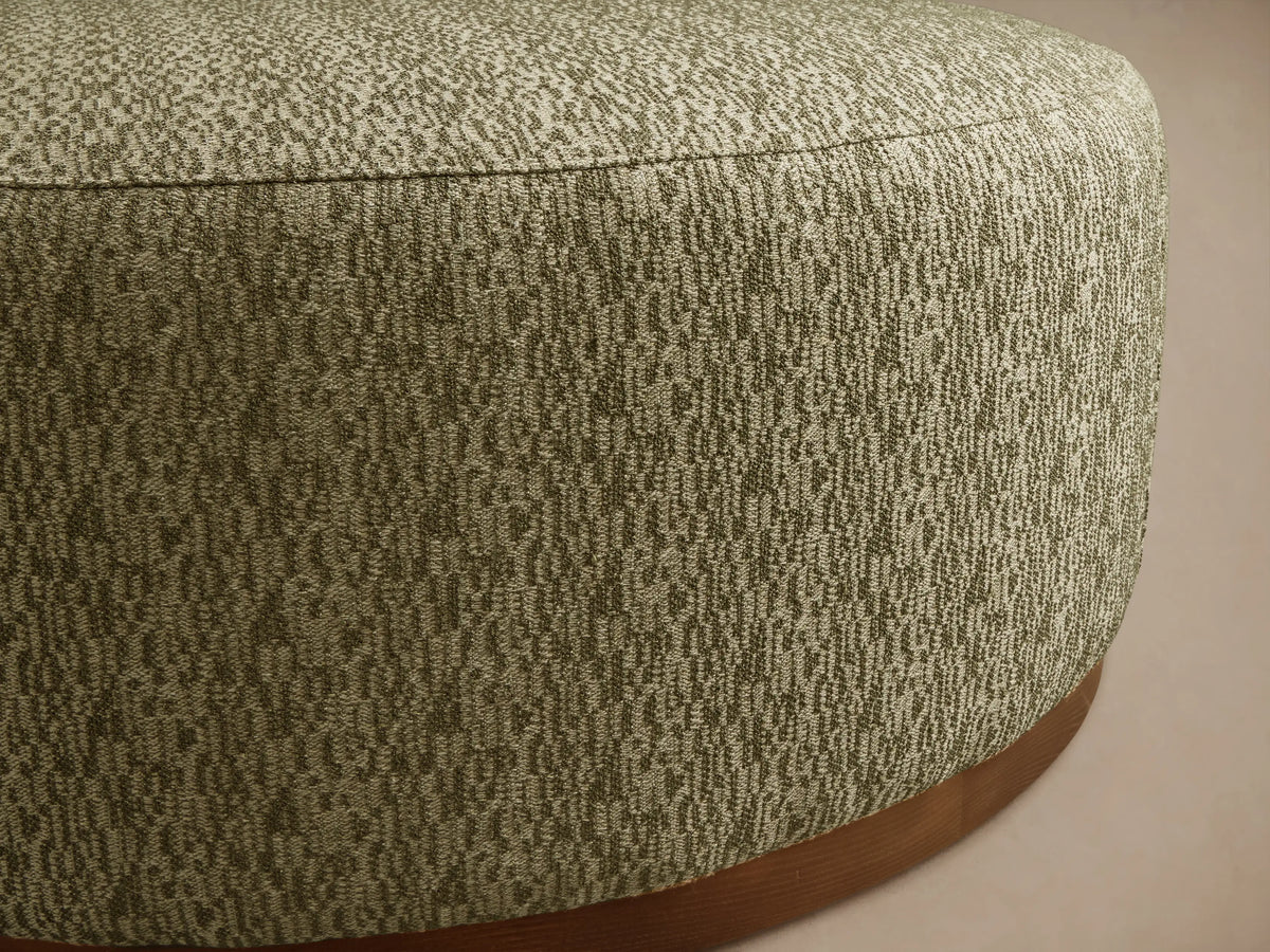 Close-up of a round Ryles Plinth Ottoman, upholstered in textured olive green and light beige flecked fabric, with a natural wood base.