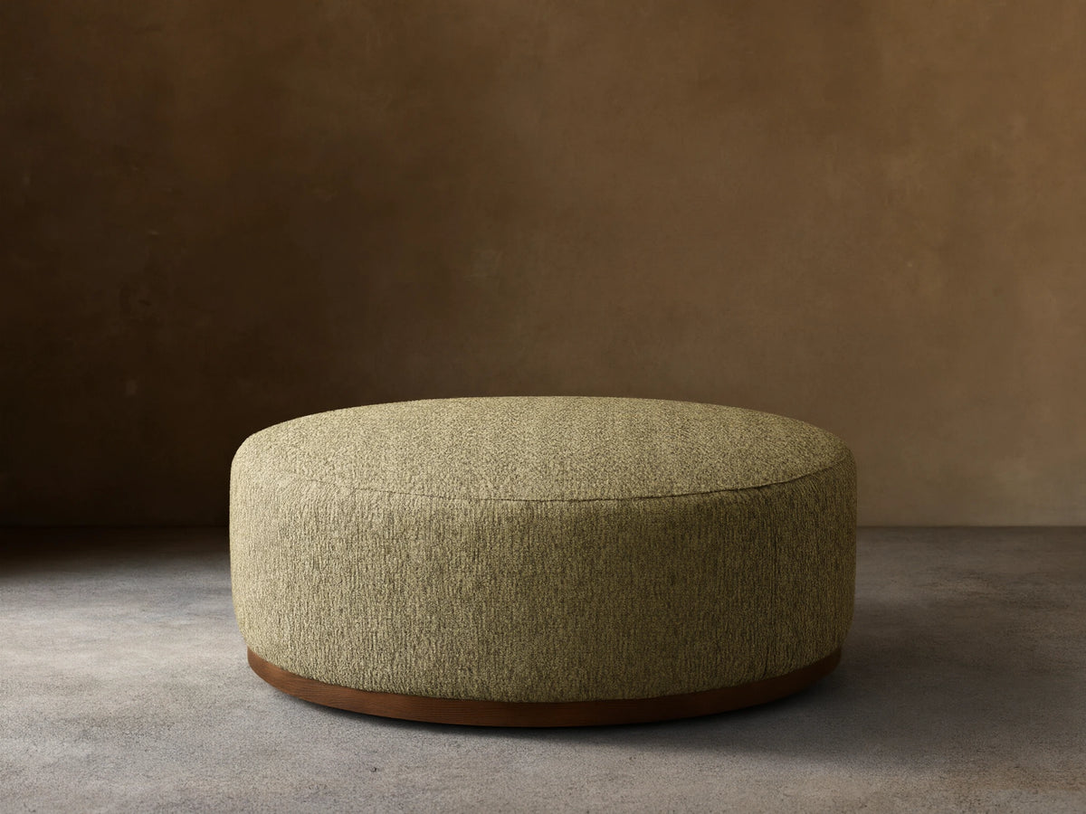 Round Ryles Plinth Ottoman, upholstered in textured light olive green fabric with a dark wood base, on a wooden floor.