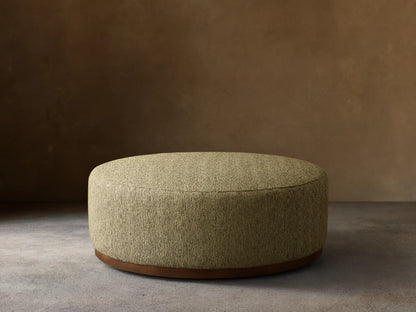 Round Ryles Plinth Ottoman, upholstered in textured light olive green fabric with a dark wood base, on a wooden floor.