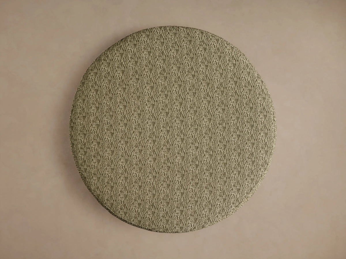 Ryles Plinth Ottoman: Round ottoman with textured olive green fabric, centered against a plain light brown wall.