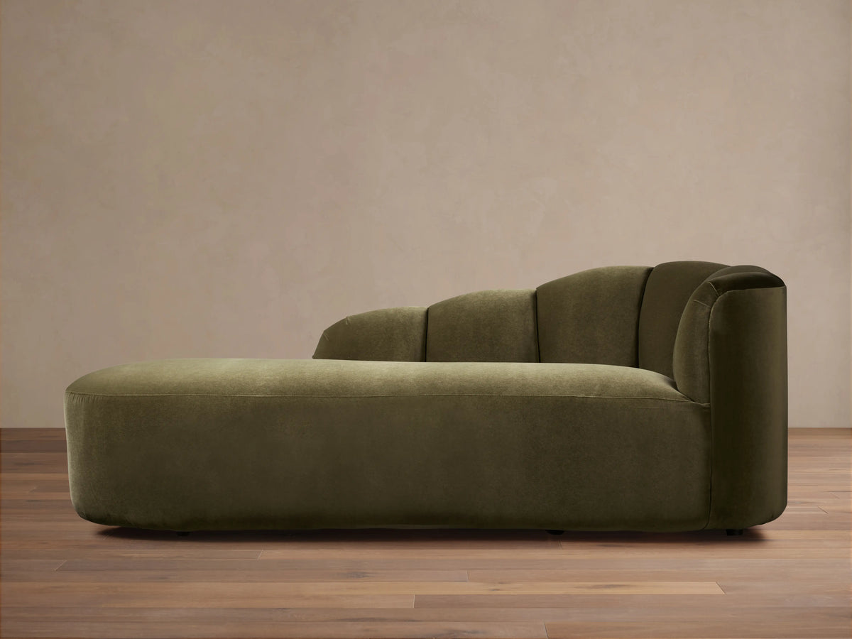 Amira Daybed: olive green velvet chaise lounge with a low, sculpturally curved backrest, on a wooden floor.