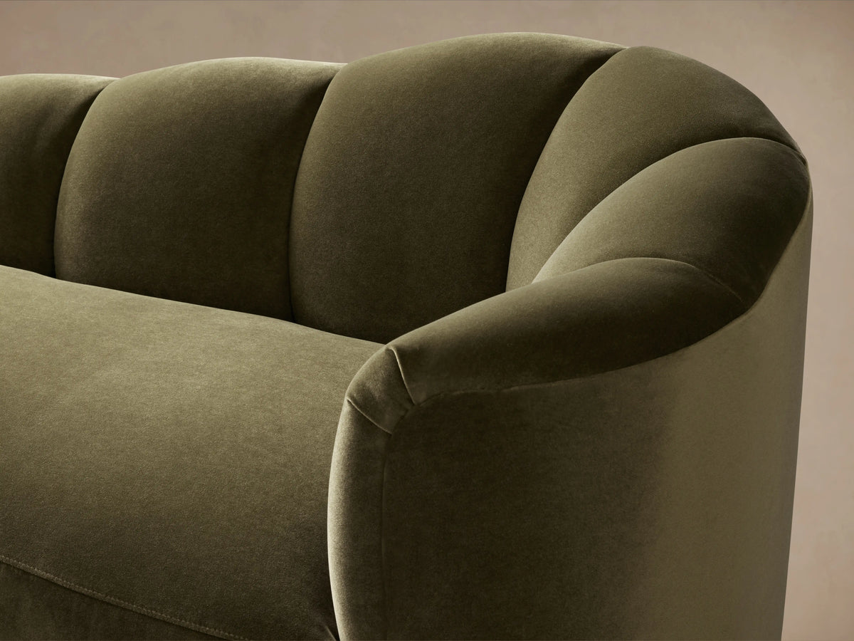 Close-up of Amira Daybed in dark olive green velvet, showing curved right arm and fluted back cushions against a soft brown wall.
