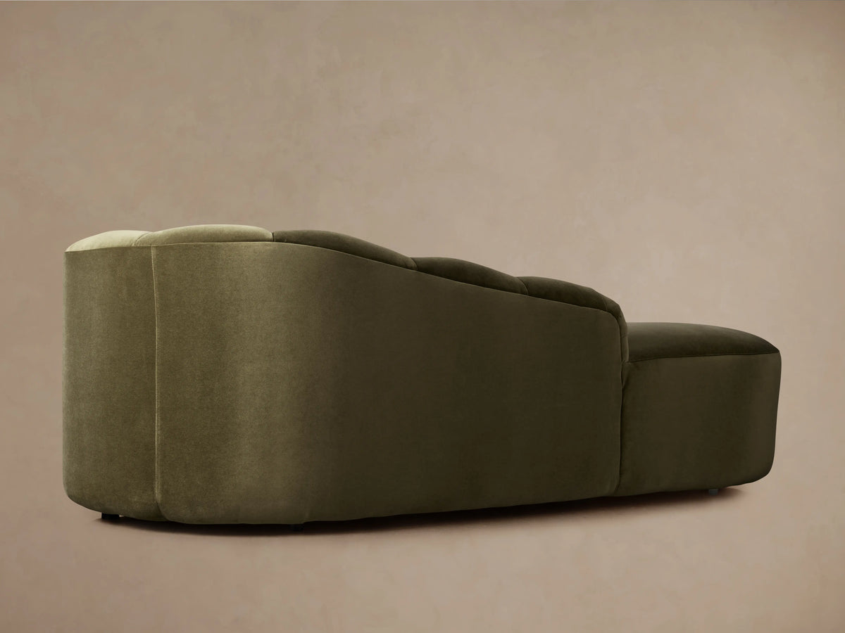 Amira Daybed: olive green velvet chaise lounge with a low, sculpturally curved backrest, on a wooden floor.